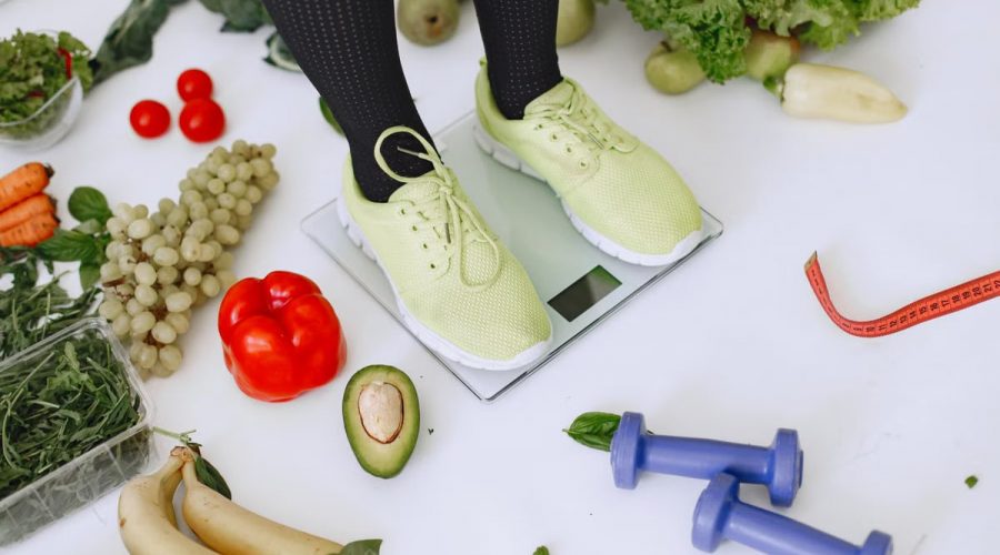 Person standing on scales surrounded by fresh produce and fitness equipment symbolizing a balanced lifestyle.